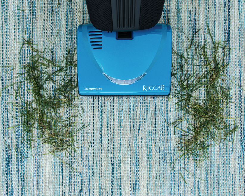 Blue Riccar vacuum cleaner cleaning a textured blue and green carpet with grass clippings.