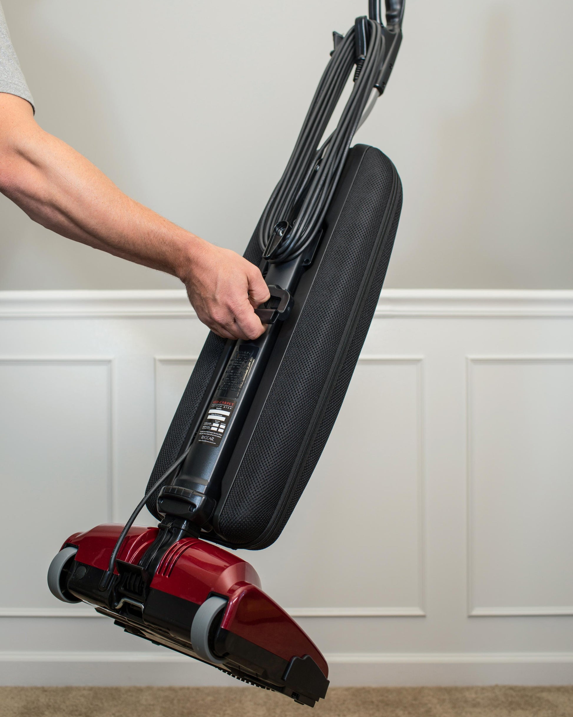 Person holding a red and black vacuum cleaner against a white wall demonstrating the easy grip carry handle on the Riccar R10P vacuum.