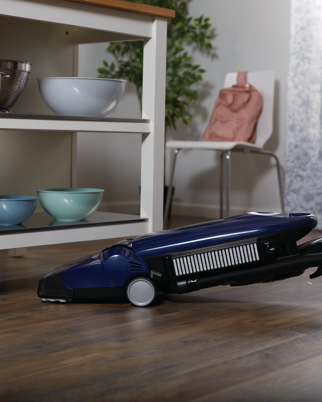 Person using a blue vacuum cleaner on a wooden floor in a room with shelves and a window.